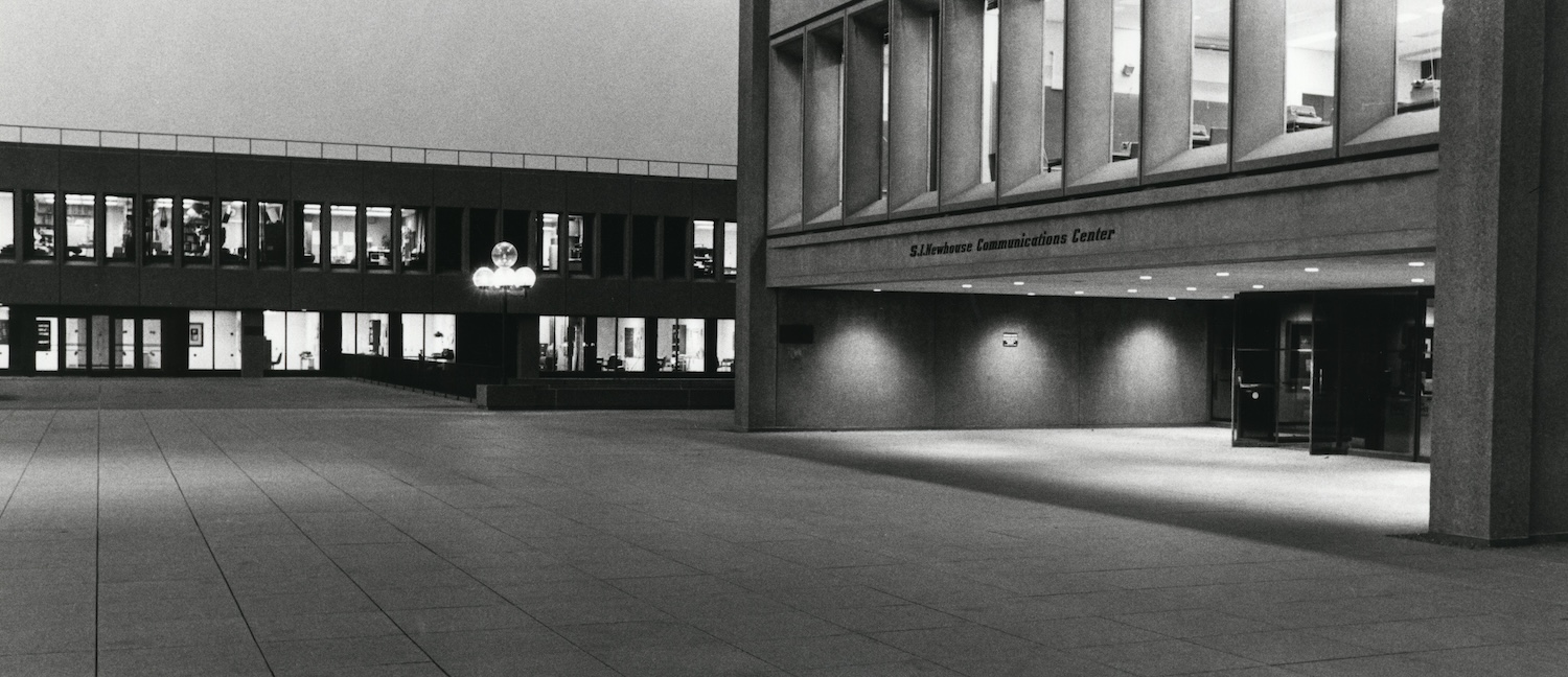 Historic black-and-white view of the S.I. Newhouse Communications Center plaza at night, with illuminated windows and an empty courtyard.
