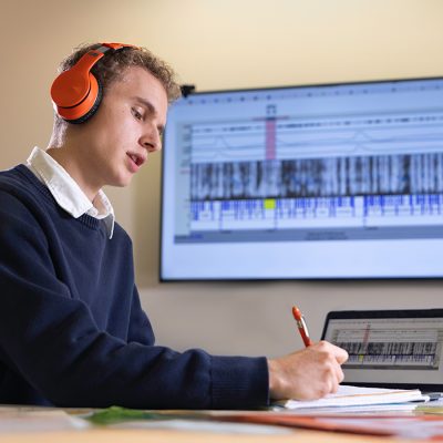 Man in orange headphones taking notes at a desk, with audio waveform and spectrogram software shown on his laptop and a larger wall-mounted screen behind him.