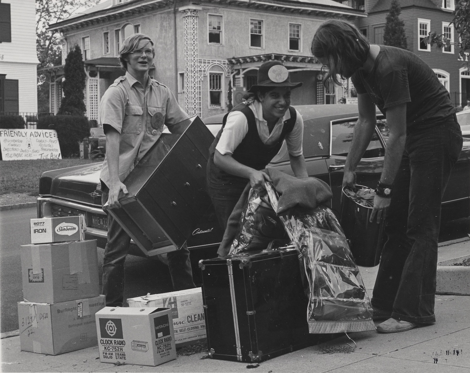 Black-and-white photo of three volunteers unloading boxes, furniture, and luggage from a car during a campus move-in.