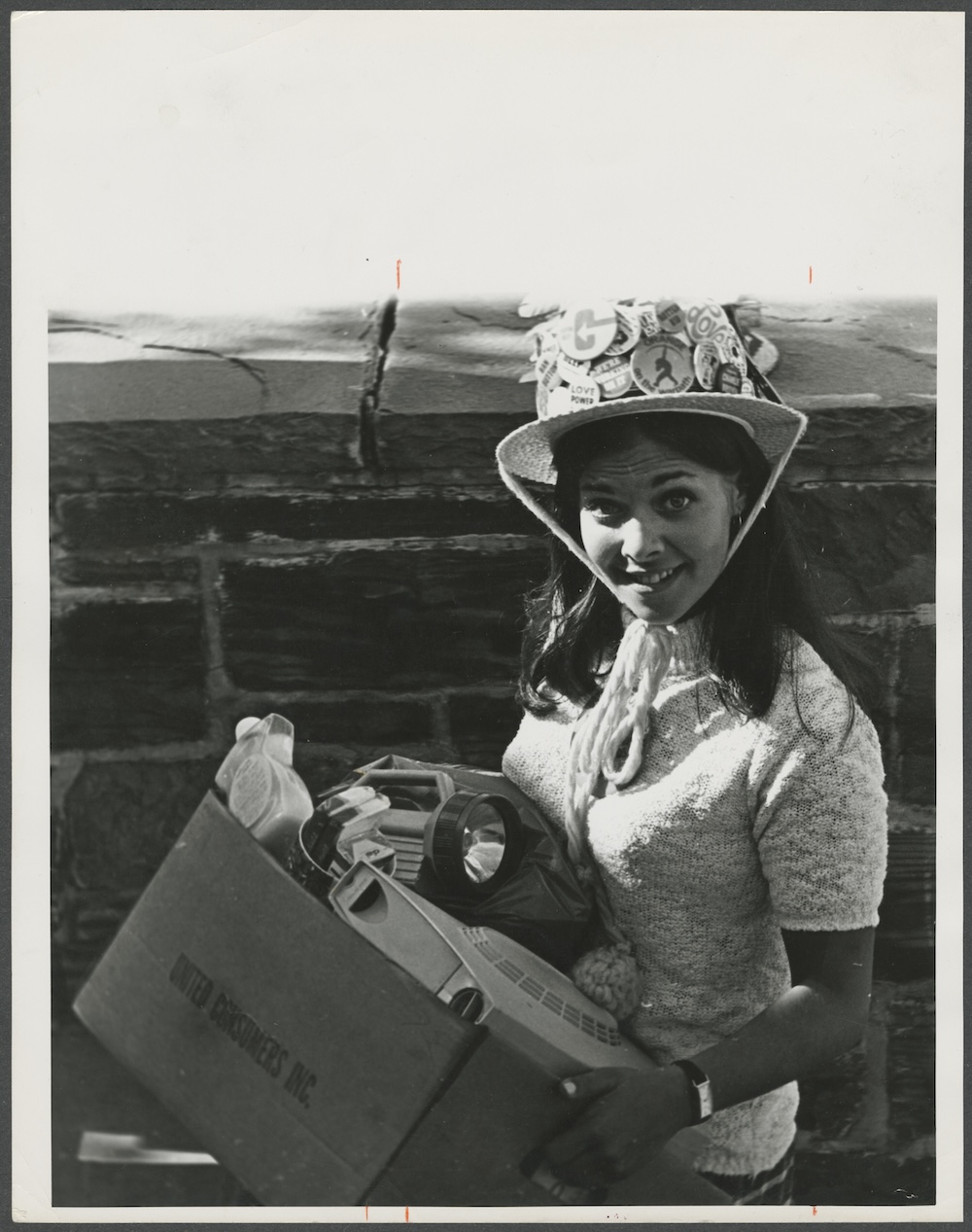Person wearing a button-covered hat carries a box of belongings, including a flashlight and projector, during a campus move-in event.