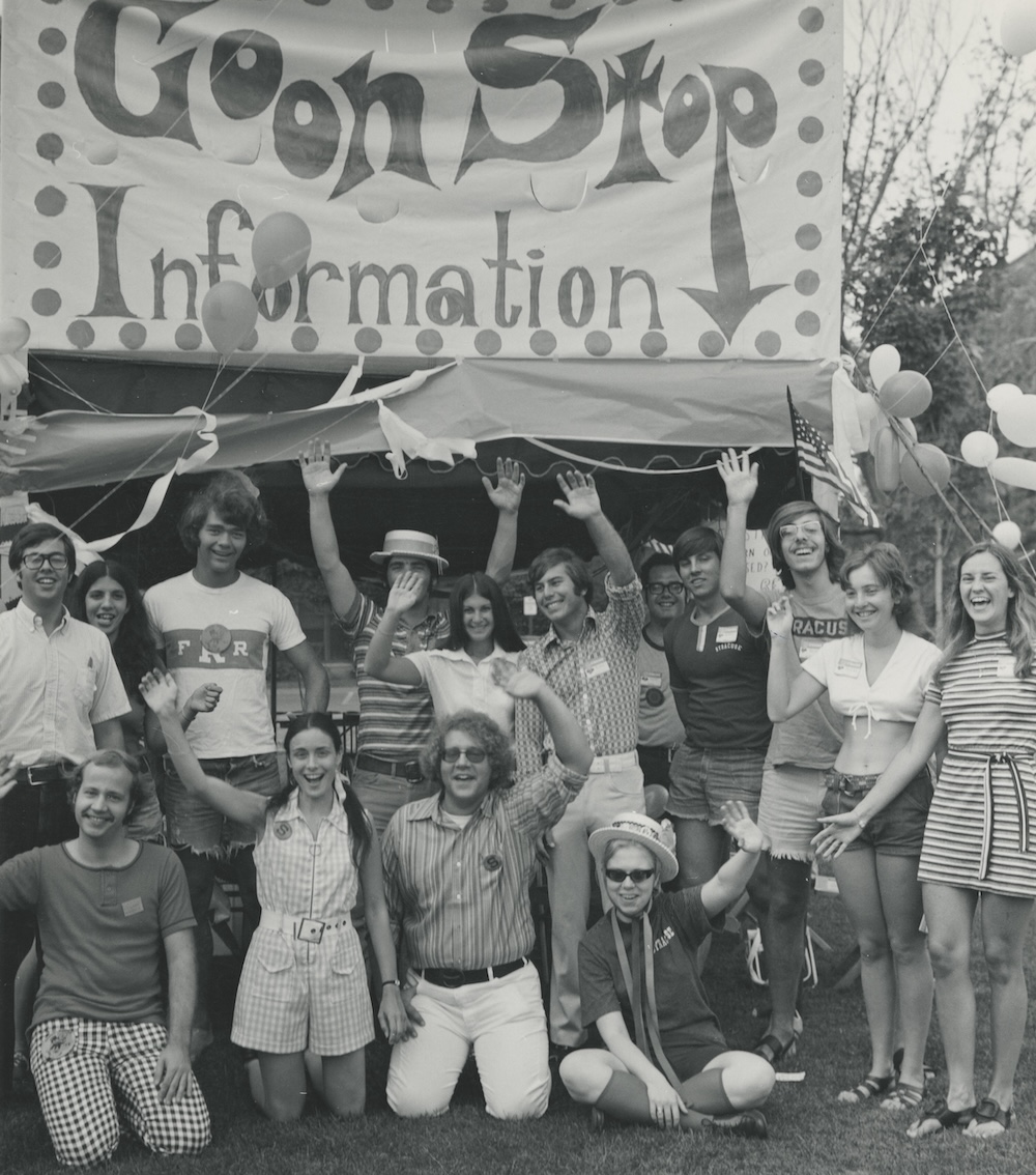Group of students gathered in front of a “Goon Stop Information” booth, waving at the camera during a campus welcome event.
