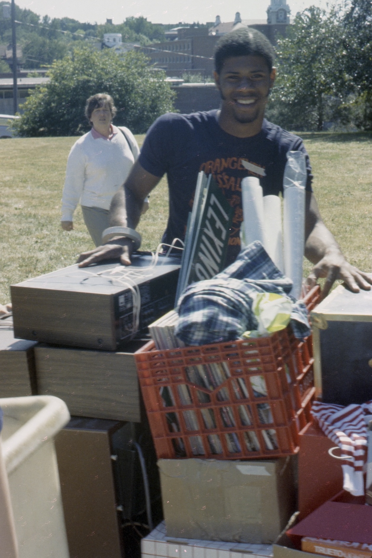 Person standing behind a pile of boxes, records, and stereo equipment outdoors during a campus move-in or collection event.
