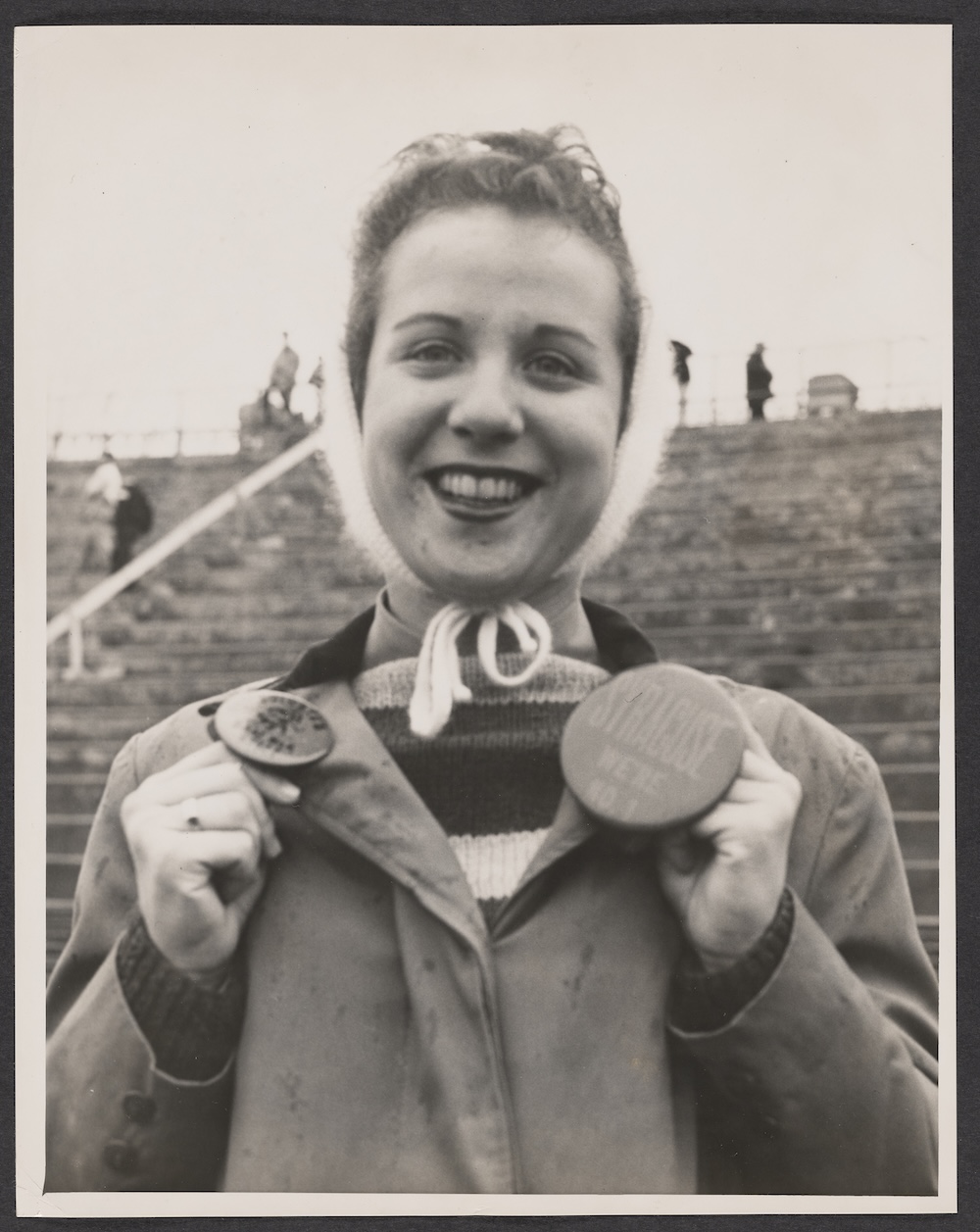 Black-and-white portrait of a person wearing a tied head covering and holding two large buttons while standing in stadium bleachers.Provide your feedback on BizChat