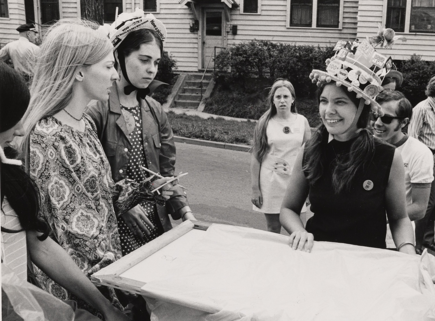 Black-and-white photo of students gathered around a large canvas during a campus welcome event, including participants wearing decorated hats.