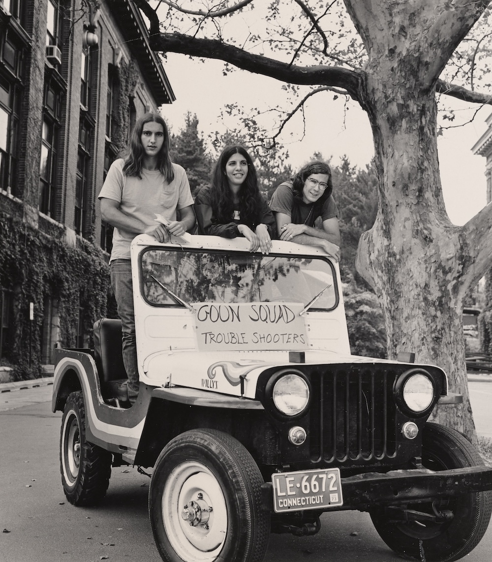 Three students pose on a Jeep with a sign reading “Goon Squad Trouble Shooters” during a campus welcome event.