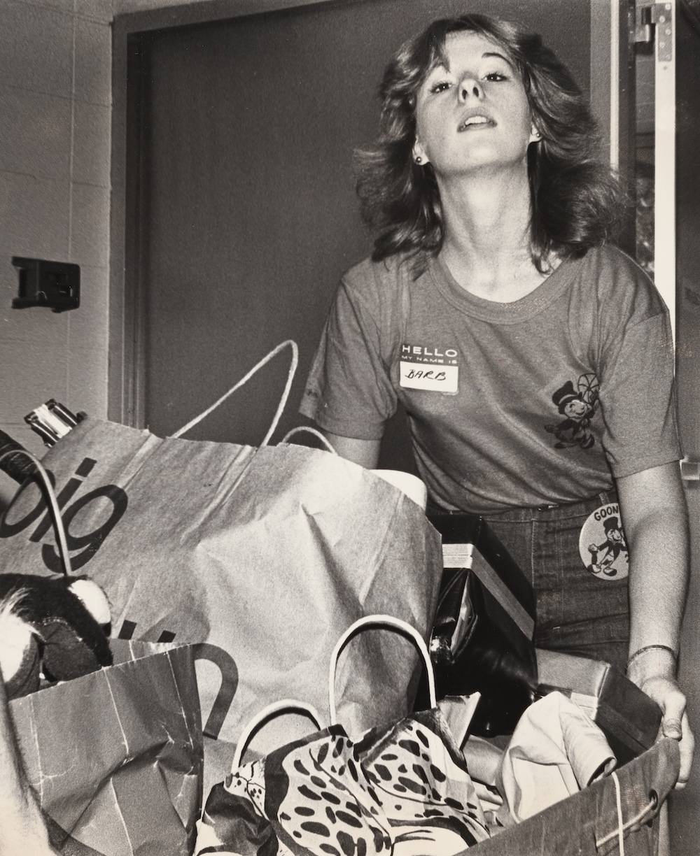 Black-and-white photo of a volunteer wearing a name tag and event button while moving bags and boxes indoors during campus move-in.