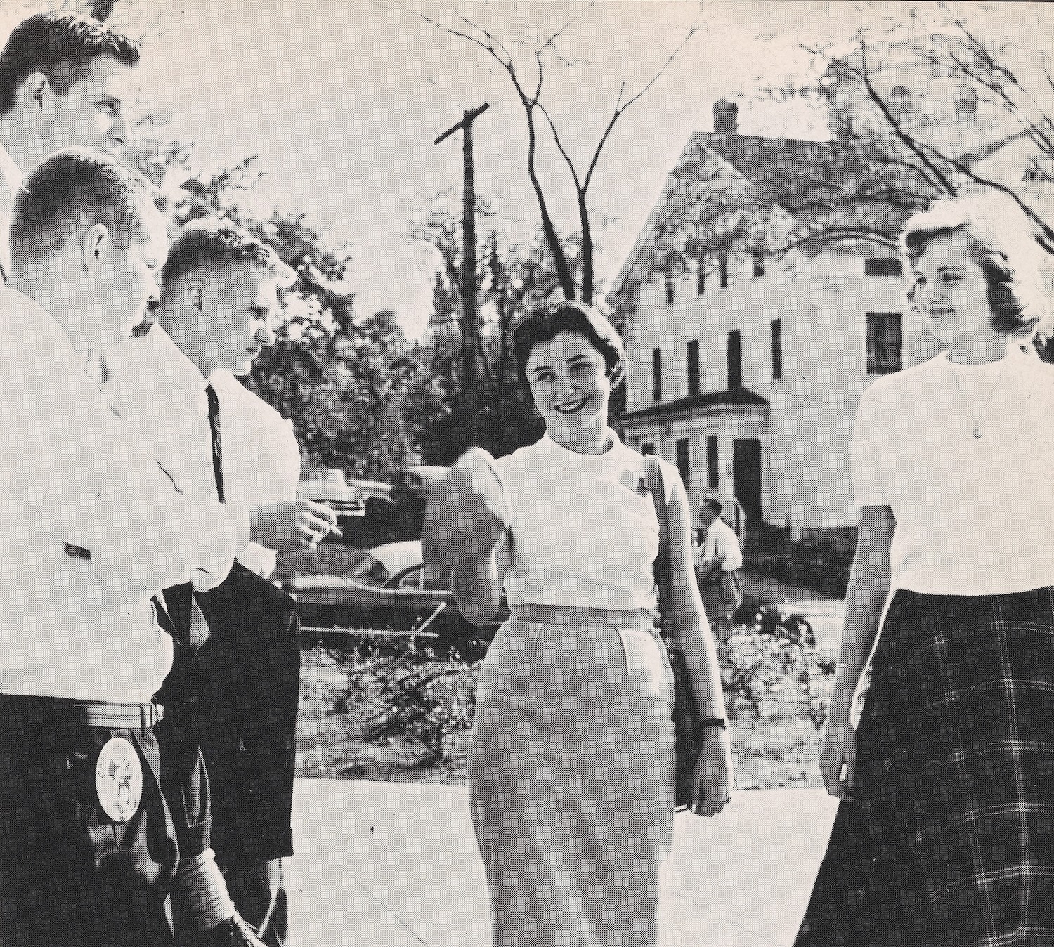 Black-and-white photo of a group of students standing and talking on a campus walkway in front of a large building.