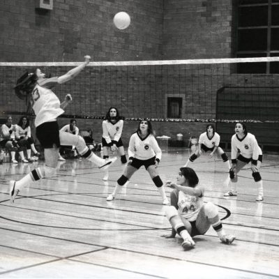 Black-and-white photo of an indoor volleyball game, with one player leaping to hit the ball over the net while teammates and opponents prepare on the court.