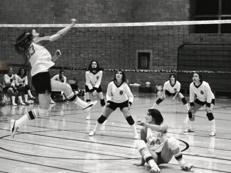 Black-and-white photo of an indoor volleyball game, with one player leaping to hit the ball over the net while teammates and opponents prepare on the court.