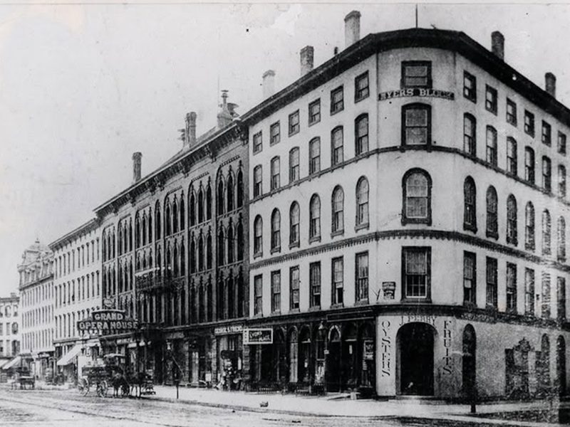 Historic black-and-white photo of a large corner commercial block with storefronts and horse-drawn carriages along a city street.