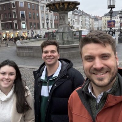 Image 1: Three people posing for a selfie in a cobblestone city square with a large fountain and historic buildings in the background.