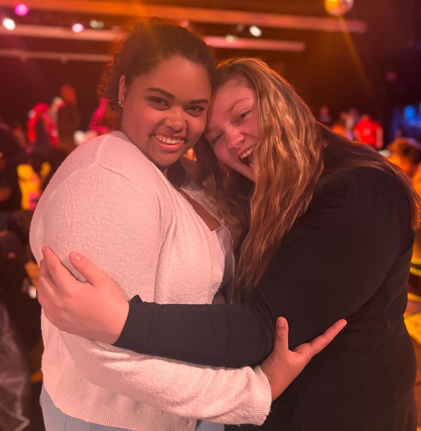 Two people embracing and posing for a photo at an indoor event with colorful stage lighting in the background.
