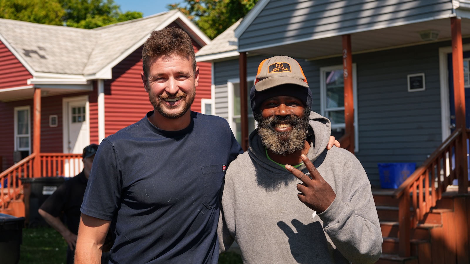 Two people pose together in front of colorful homes in a residential neighborhood.