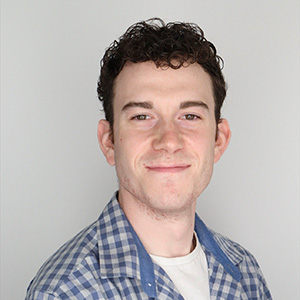 Professional headshot of a smiling man with curly dark hair, wearing a blue and white plaid shirt, against a plain light gray background.