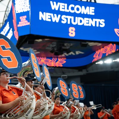 Marching Band sousaphones play beneath a "Welcome New Students" screen.