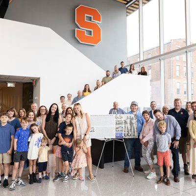Family group photo of about 30 people spanning several generations, gathered on a staircase inside a Syracuse University building beneath an orange "S" logo. A memorial poster honoring "William 'Bill' Byrne" stands on an easel in the foreground.