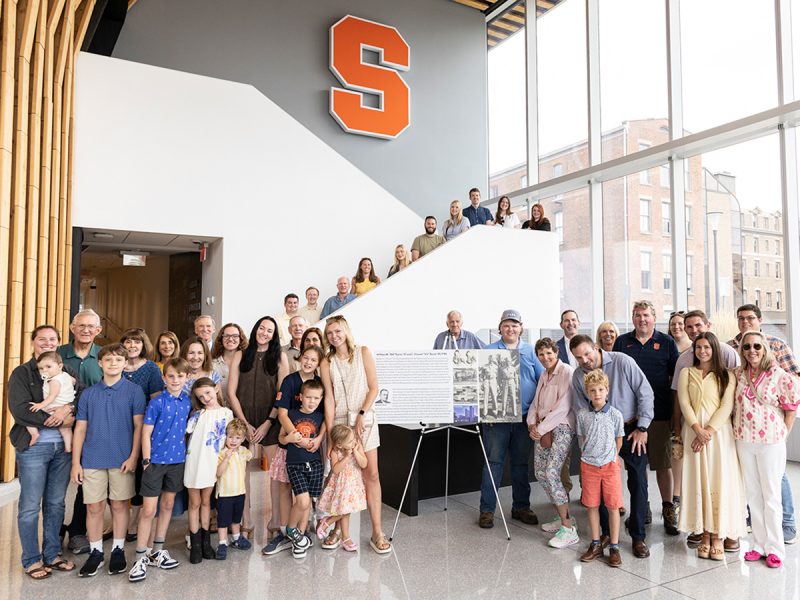 Family group photo of about 30 people spanning several generations, gathered on a staircase inside a Syracuse University building beneath an orange "S" logo. A memorial poster honoring "William 'Bill' Byrne" stands on an easel in the foreground.