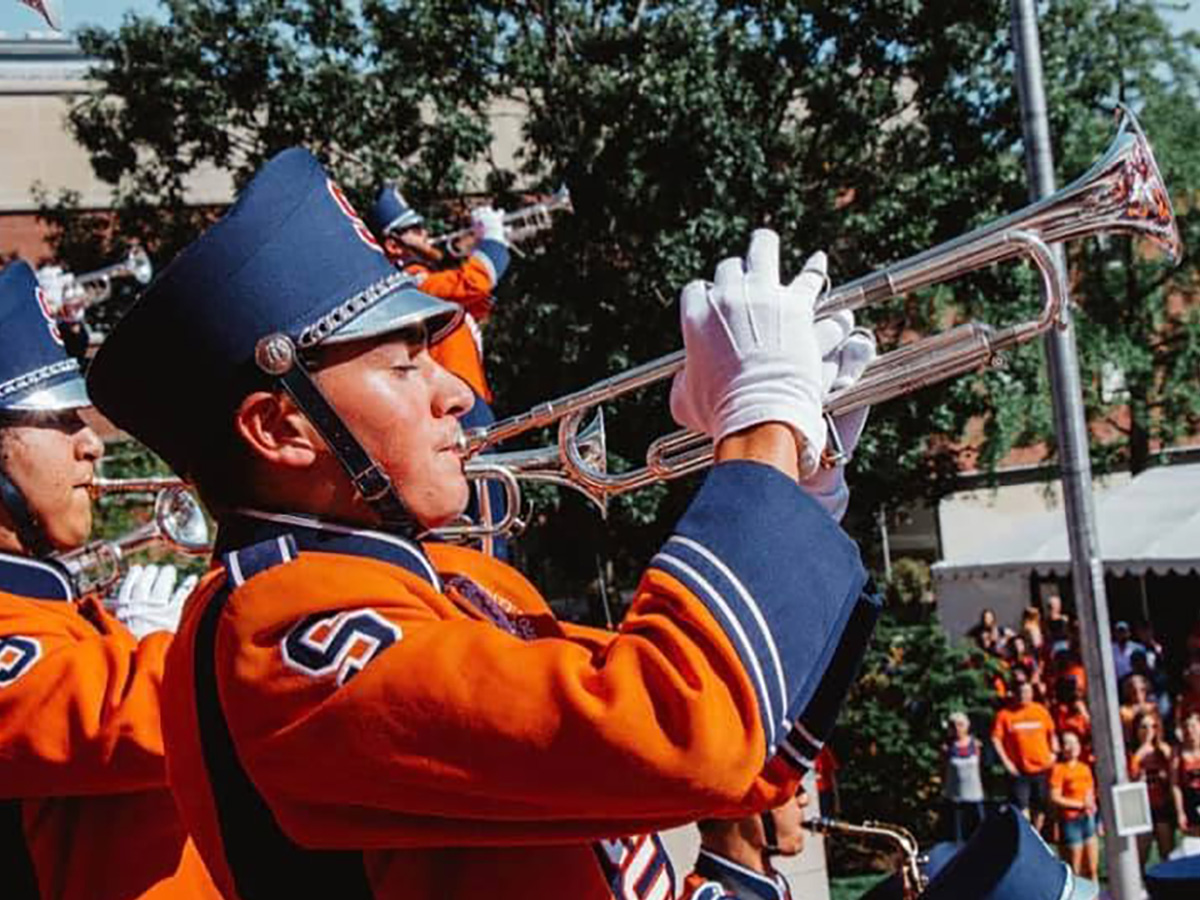 Close-up of a Syracuse University marching band trumpet player in an orange and navy uniform with white gloves, performing outdoors with bandmates and spectators visible in the background.