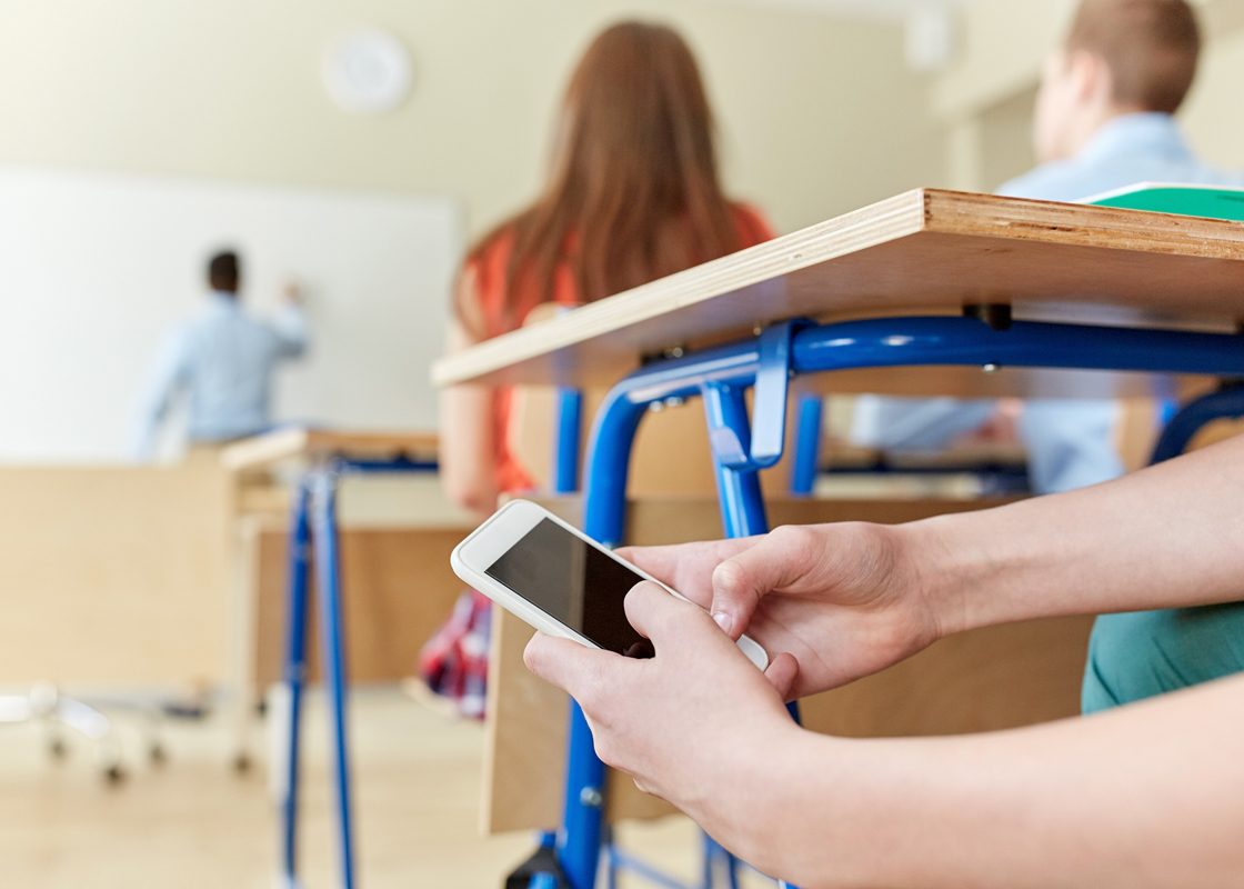 Student secretly checks smartphone under desk during class, other students in background.