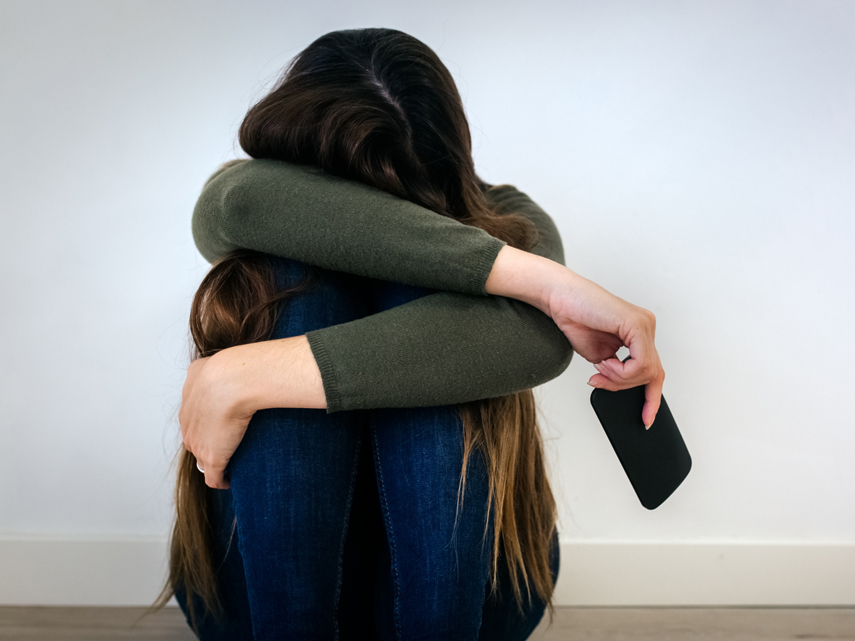 Woman sits with head buried in arms, holding smartphone at her side.