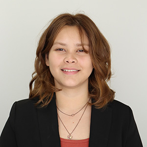 Professional headshot of a smiling woman with shoulder-length wavy auburn hair, wearing a black blazer with a necklace, against a plain light gray background.