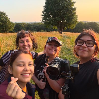 Four friends smiling together in a golden, sunlit meadow at sunset, with tall grass and trees in the background. One holds a Canon camera with a large lens mounted on a rig, suggesting they're on a filmmaking outing.