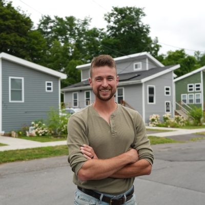 Person standing with arms crossed on a residential street, with newly built homes in the background.