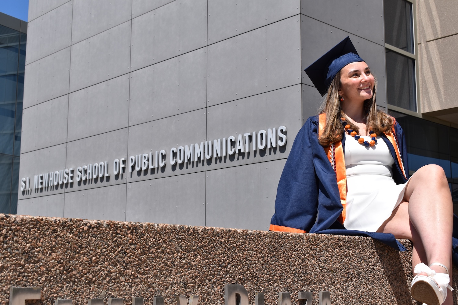 Graduate in cap and gown seated outside the S.I. Newhouse School of Public Communications.