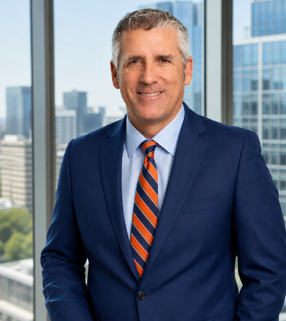 Professional headshot of a person in a blue suit and striped orange-and-blue tie, standing in an office with city skyline windows in the background.