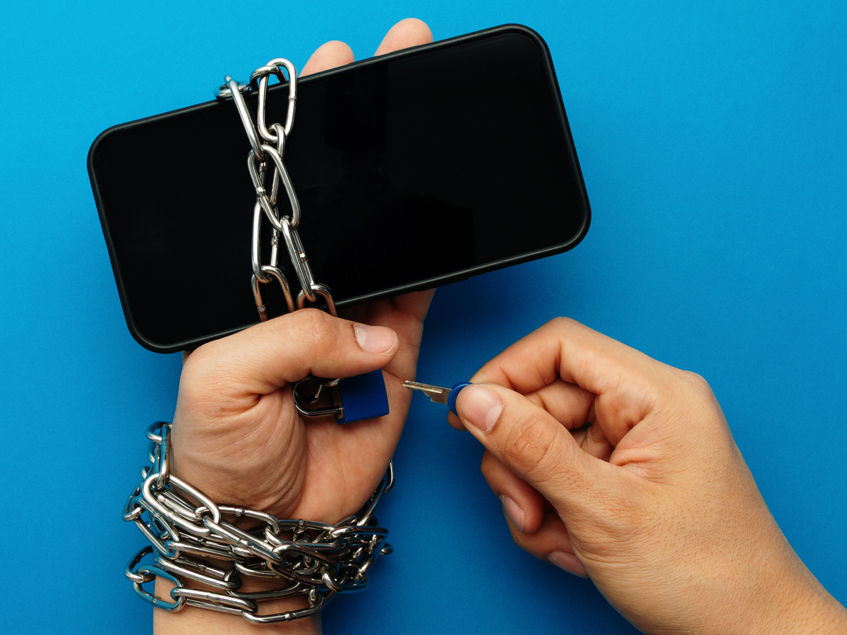 Hands chained to smartphone with padlock, one hand holding a key