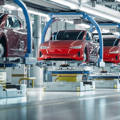 Row of red electric vehicle bodies suspended on automated guided vehicles (AGVs) along a modern factory assembly line, with blue overhead robotic arms and bright overhead lighting in a clean, high-tech manufacturing facility.