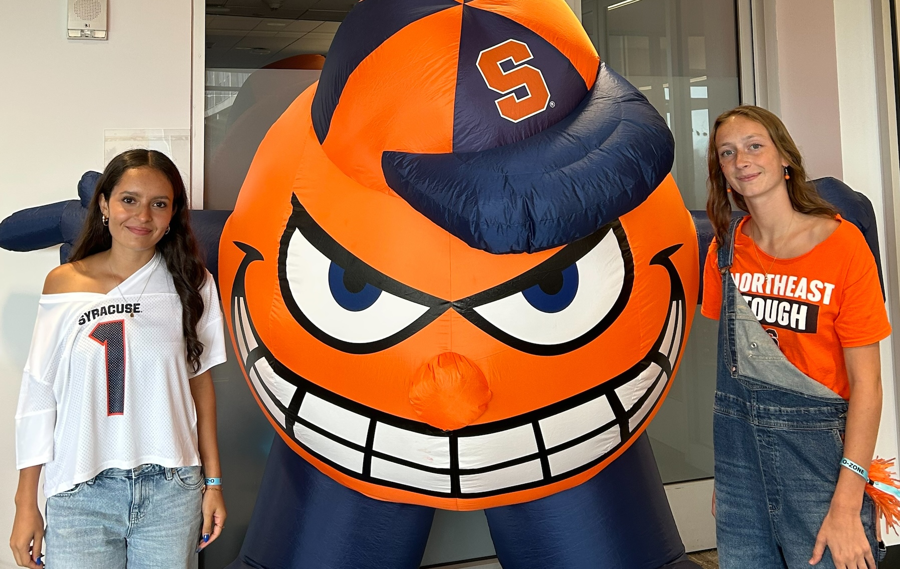 Two people standing beside a large orange Syracuse University mascot display indoors.