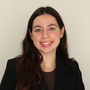 Professional headshot of a smiling woman with long dark hair, wearing a dark blazer, against a plain light gray background.