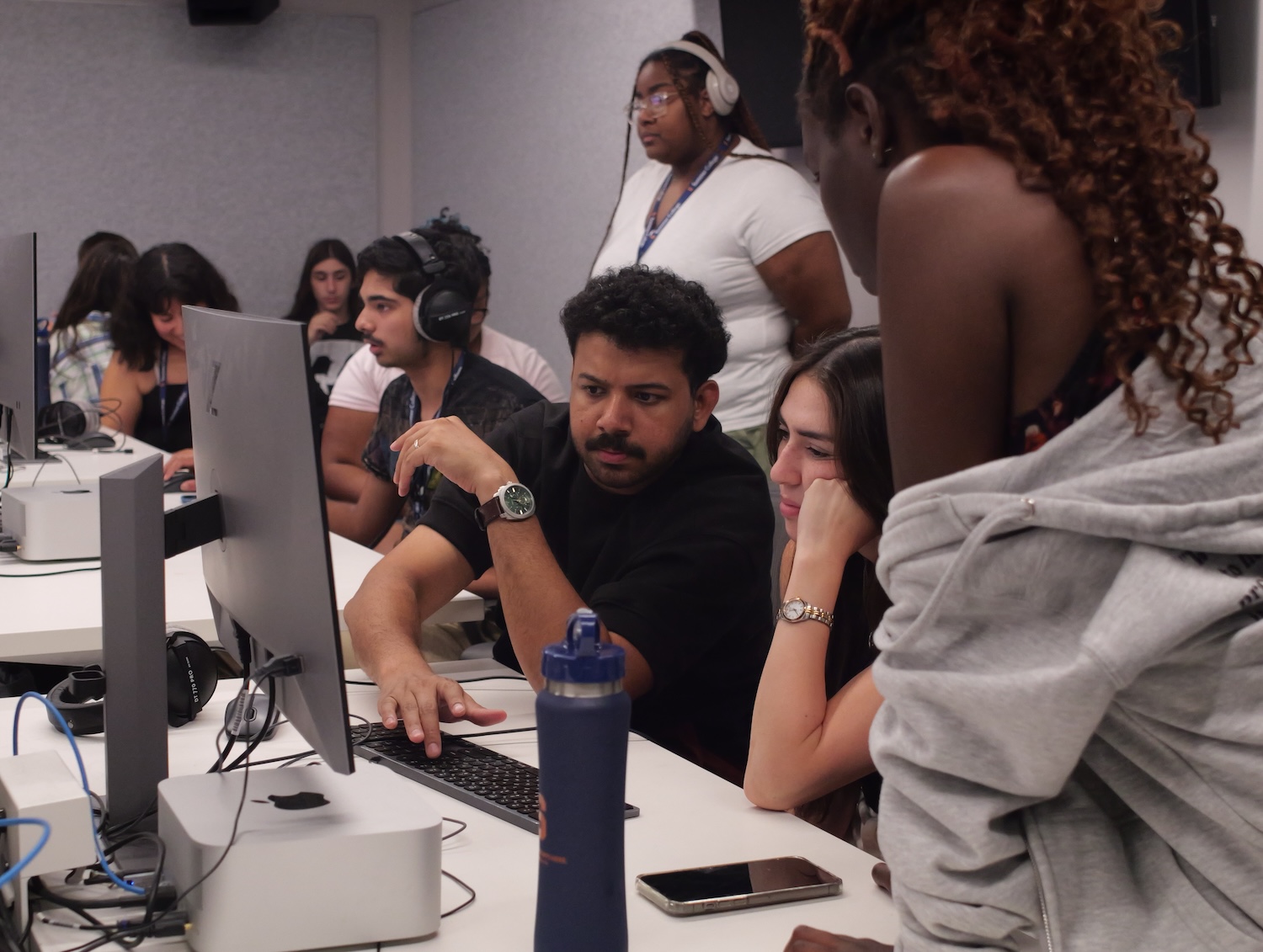 Students collaborate around desktop computers in a digital media lab, reviewing work on a shared screen.