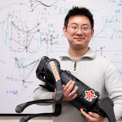 Person holding a quadruped robot in front of a whiteboard covered with technical diagrams and equations.