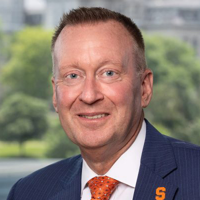 Headshot of a person in a navy suit and orange tie with an orange 'S' pin, campus greenery blurred in the background