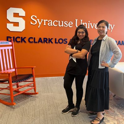 Two women pose beside an orange rocking chair in front of an orange wall reading "Syracuse University Dick Clark Los Angeles Program," with a white couch nearby.