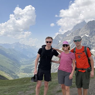 Three people pose on a mountain trail with jagged peaks behind them.