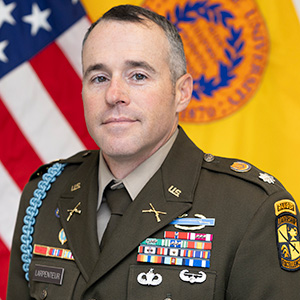 Portrait of a U.S. Army lieutenant colonel in a green service uniform with ribbons, badges, and rank insignia, standing before an American flag and a yellow branch flag.