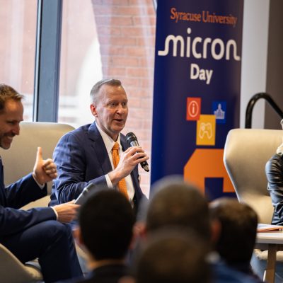 Person speaking into a microphone on a panel beside "Syracuse University Micron Day" banner.