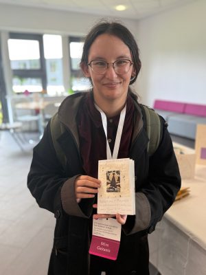 A person holds up a handmade zine titled "Nothing About US Without US: Reflections & Musings," made for the 17th International Disability Law Summer School, June 2026. Their conference badge reading "Mira Gaitanis" hangs below it.