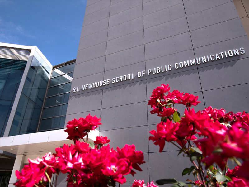 Exterior of the S.I. Newhouse School of Public Communications building, with red roses in the foreground and a clear blue sky above.