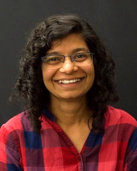 Headshot of a smiling woman with curly hair and glasses, wearing a red and blue plaid shirt, against a dark background