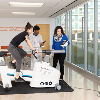 A person pedals a stationary exercise bike while two colleagues observe nearby, in a research space near a wall reading 'Nutrition Assessment, Consultation & Education (ACE) Center II.'