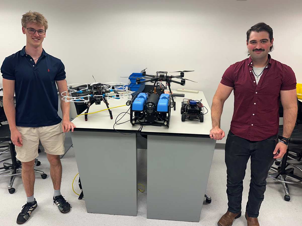 Two students pose beside drones and a submersible robot on display tables.