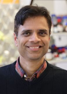 Pranav Soman smiles at the camera while standing in a lab, wearing a black sweater over a plaid shirt, with shelves of lab supplies and equipment visible behind him.