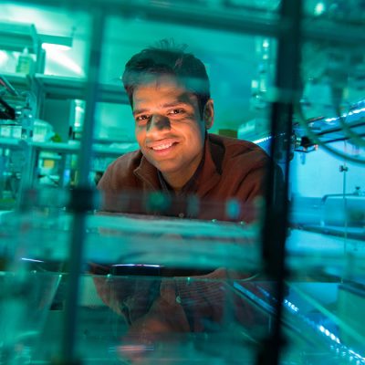 Pranav Soman smiles from behind lab equipment bathed in blue-green light, surrounded by glass panels, tubing, and machinery in a research lab.