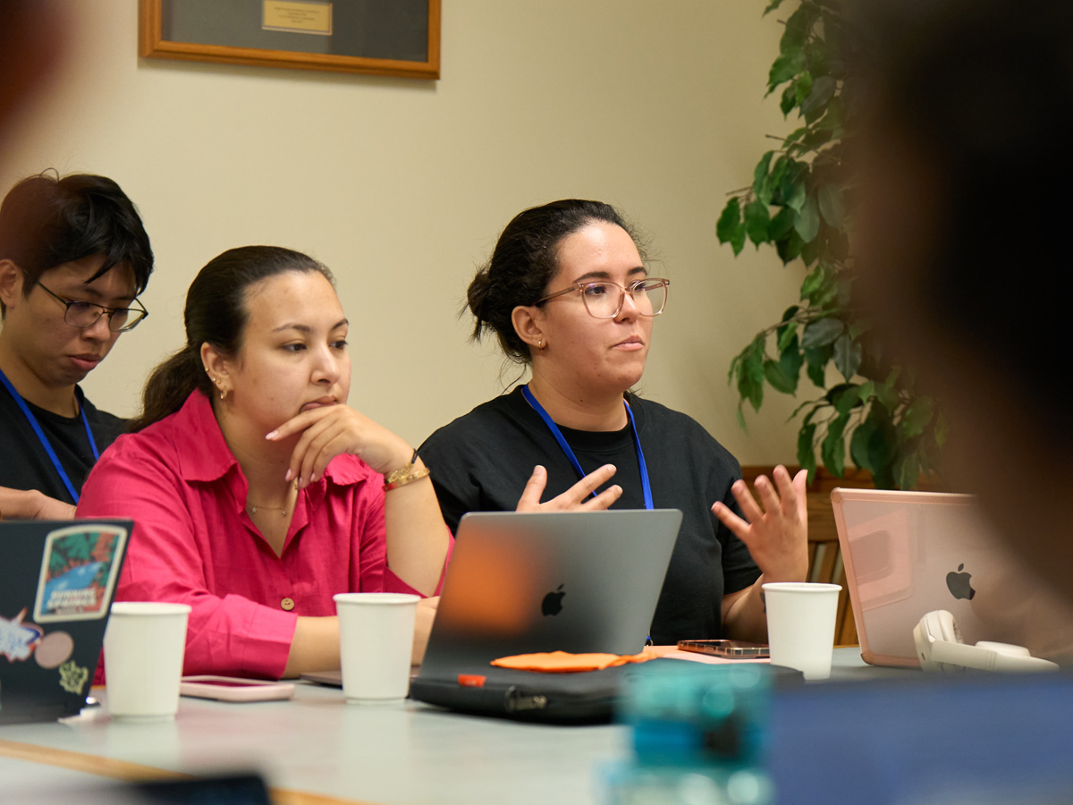 Three researchers at a table with laptops, one gesturing while speaking