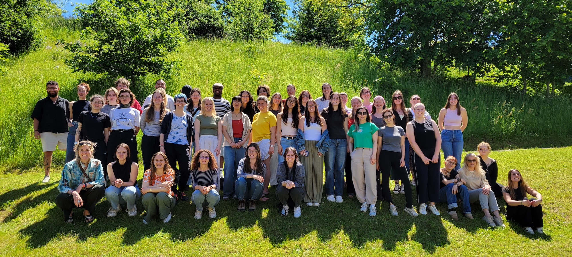 Large group photo taken outdoors on a sunny day, with participants standing and sitting on a grassy hillside surrounded by trees.