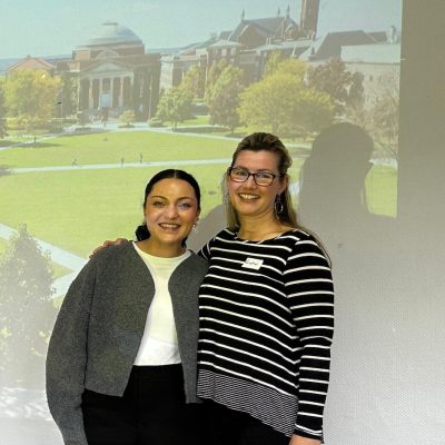 Two presenters stand in front of a projected slide.